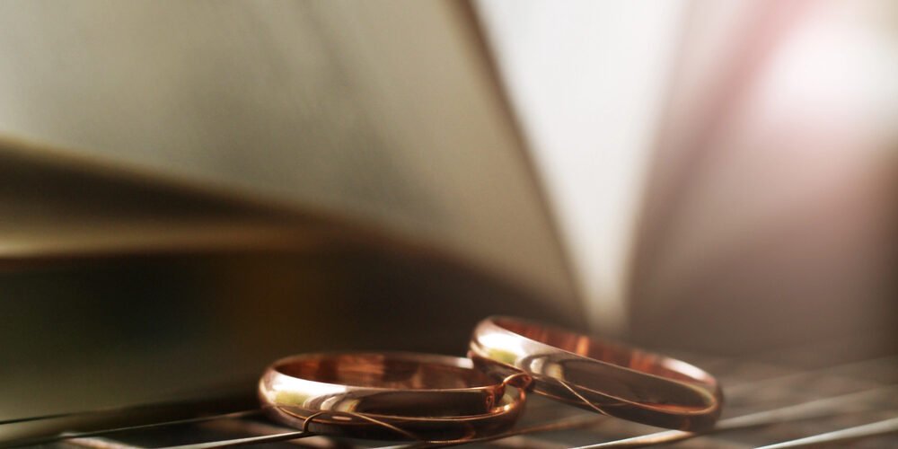 Two gold wedding rings placed on a reflective surface with a blurred open book in the background, symbolizing divorce and legal strategies.